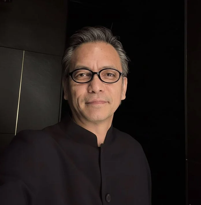 グレーヘアと眼鏡をかけた男性が、黒い上着を着用し、穏やかな笑顔を浮かべているポートレート画像です。暗い室内で撮影されています。