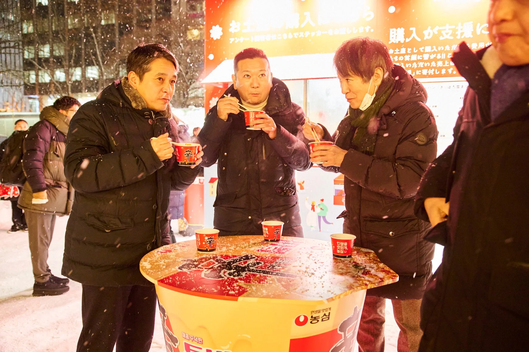 雪が降る寒い夜、屋外イベントで数人の男性が「辛ラーメン」のカップ麺を食べて温まっている様子です。