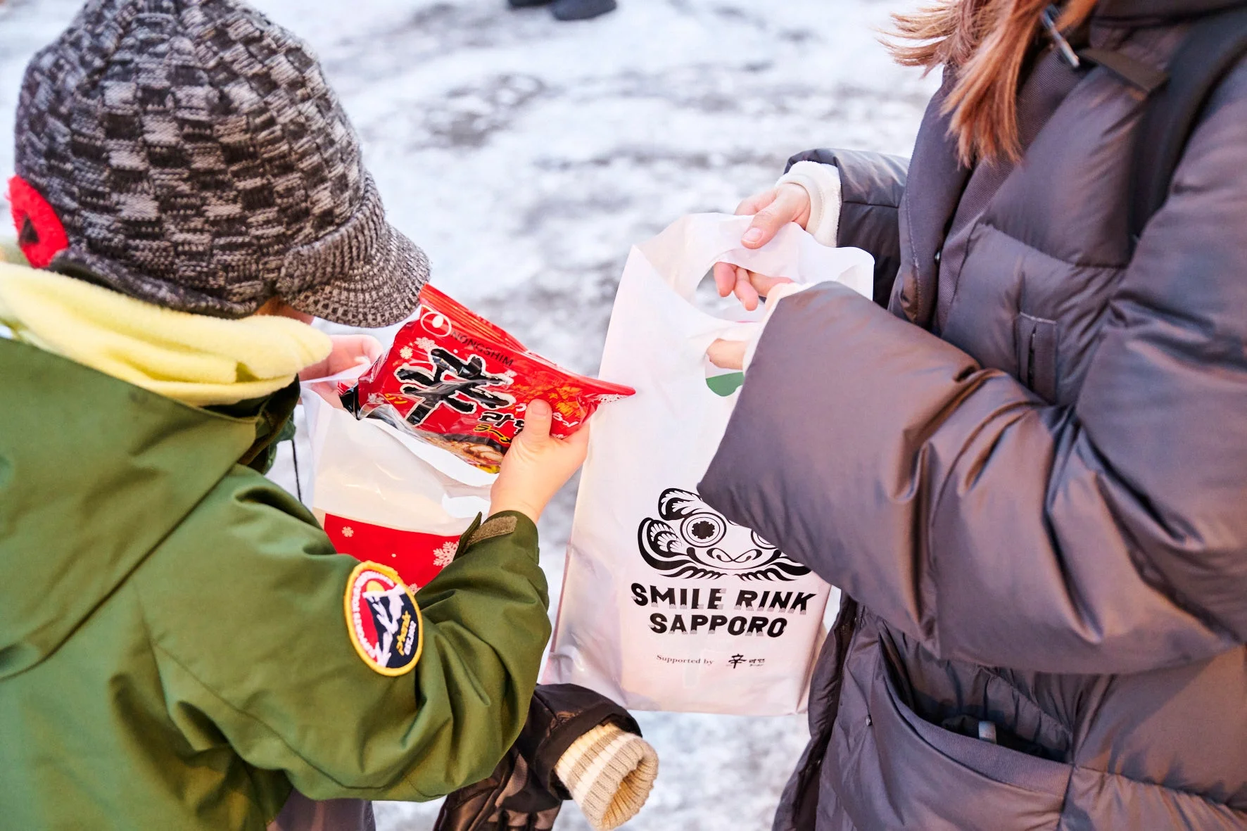 雪景色の中、防寒着を着た子供が大人からインスタントラーメンの袋を受け取っている。