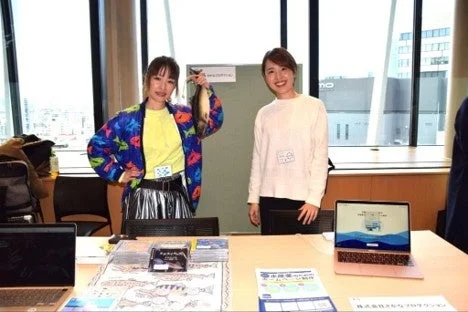 オフィスのような場所で、二人の女性が魚を持ち、笑顔を見せている