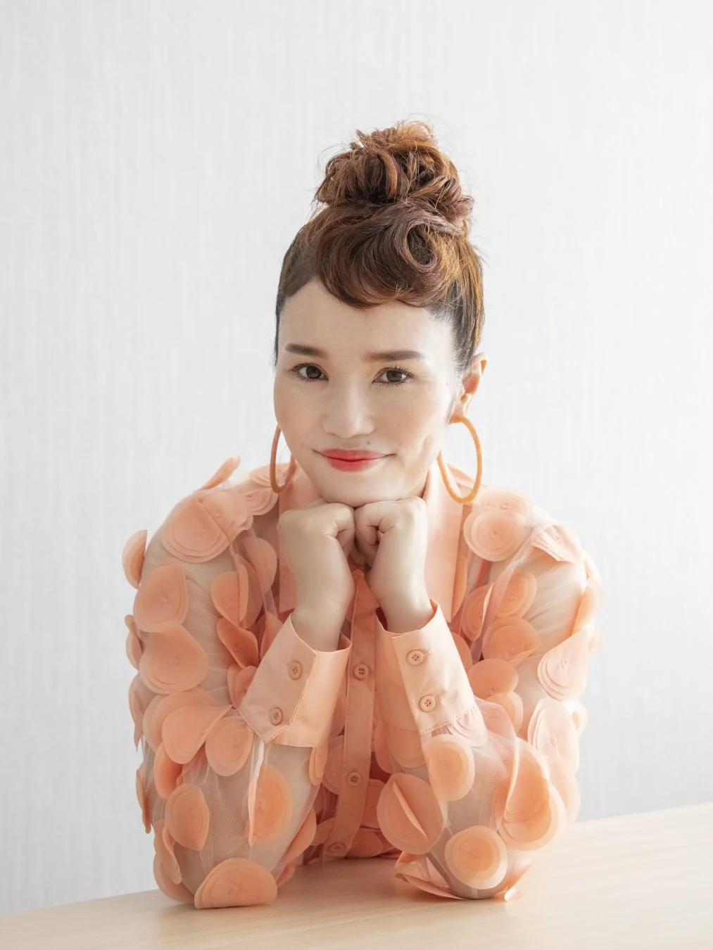 ピーチカラーの個性的なブラウスを着た女性が、お団子ヘアで笑顔を見せています