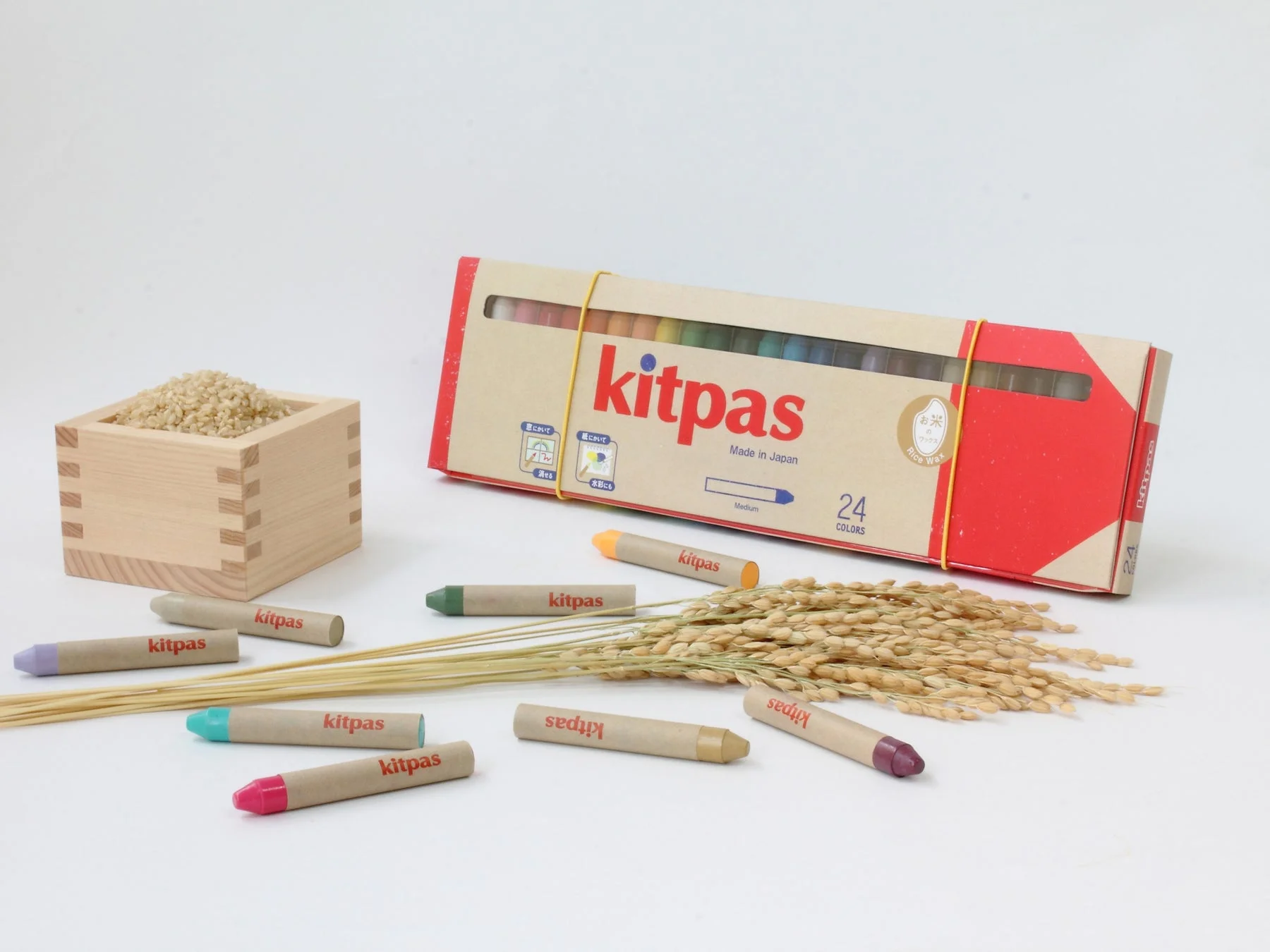 お米のワックスで作られた日本製クレヨン「kitpas」のセットと、その原料である米（玄米と稲穂）が並べられた写真