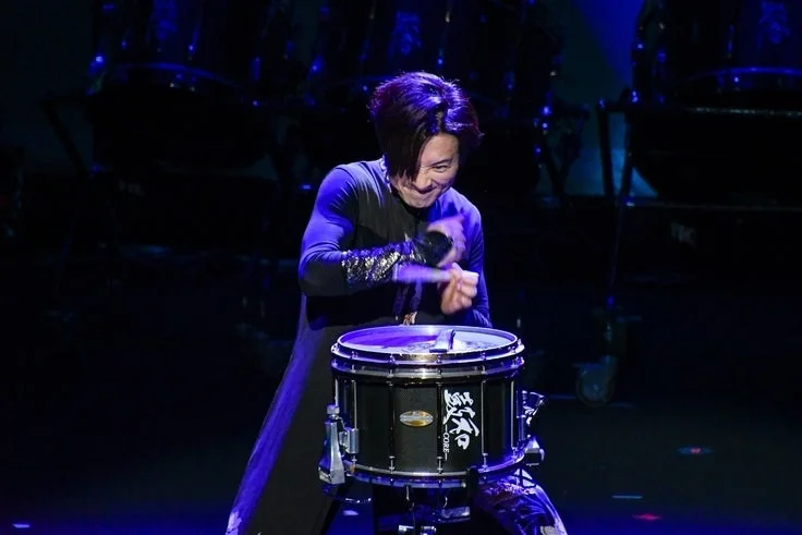 暗いステージ上で、青い照明を浴びたパフォーマーがスネアドラムを激しく演奏しています