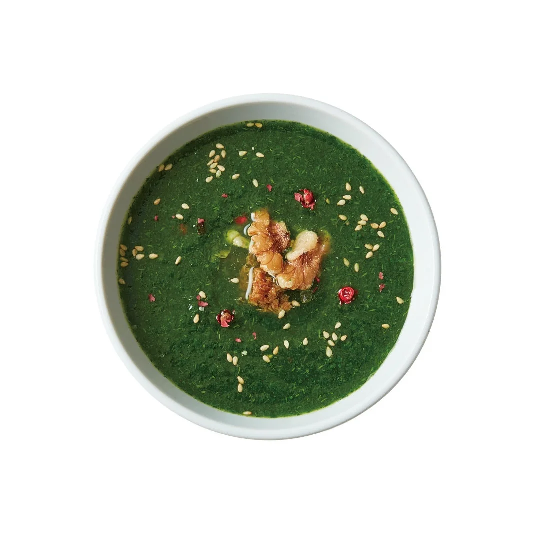 白いボウルに盛られた鮮やかな緑色のスープです。表面には香ばしいごまが散りばめられ、中央には食感のアクセントとなるくるみが添えられています。赤色の粒が彩りを添え、健康的で美しい一皿です。
