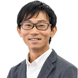 眼鏡をかけたアジア系の男性が、白いシャツとグレーのジャケットを着用し、笑顔でカメラを見ている上半身のポートレートです。