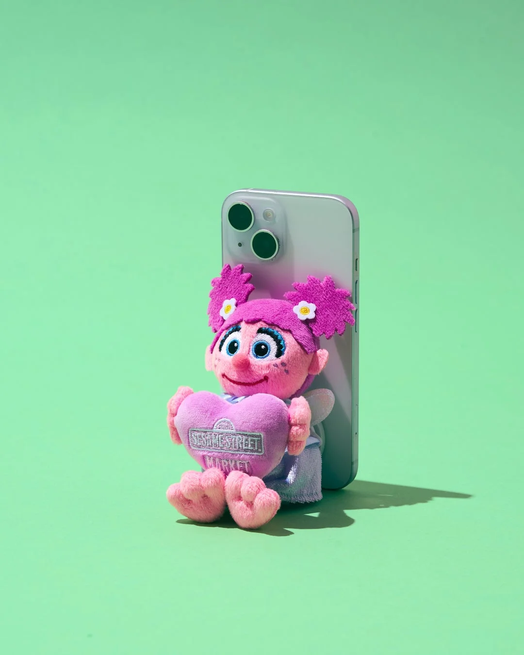 セサミストリートのアビー・カダビーのぬいぐるみが、ピンクのハートを抱えて座っており、その背後にはシルバーのスマートフォンが立てかけられています