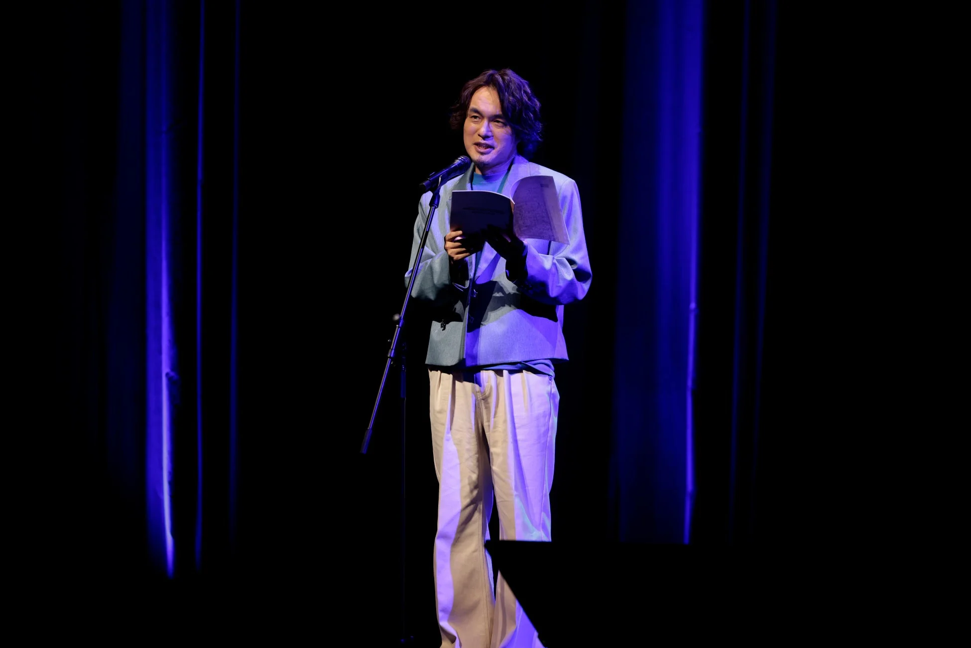 青い照明のステージでマイクを持つ男性の姿