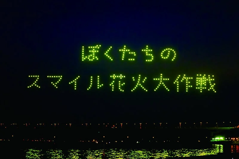 夜空に多数のドローンが光で「ぼくたちの スマイル花火大作戦」という日本語のメッセージを形成している様子。水面には街の明かりが反射し、幻想的な夜のイベント風景です。