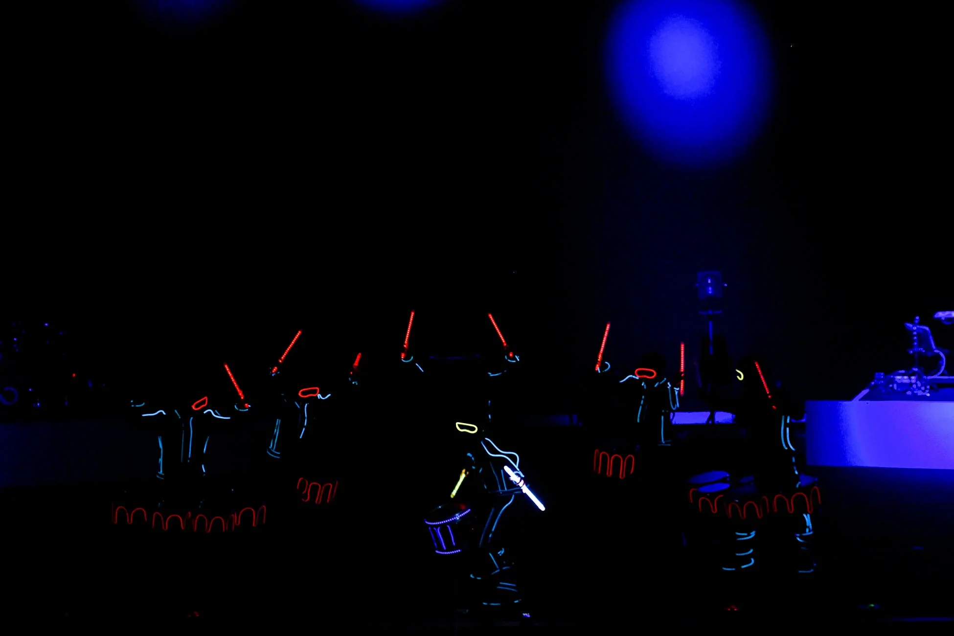 暗闇のステージで、ELワイヤーで光る衣装をまとったパフォーマーたちがドラムを演奏している様子