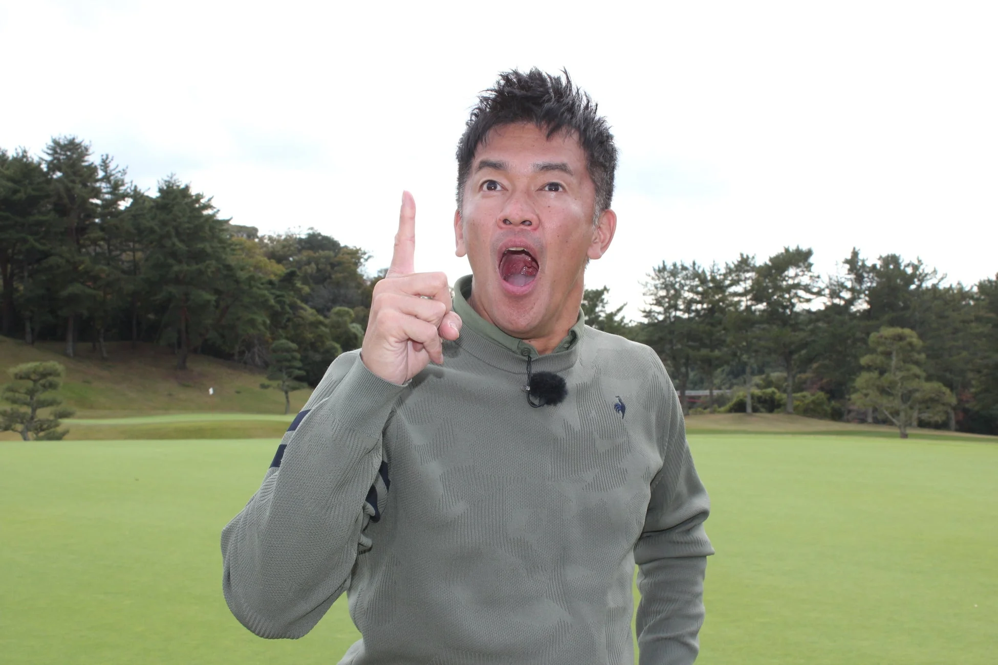 ゴルフ場に立つ男性が、口を大きく開けて驚いた表情で、人差し指を上に向けている