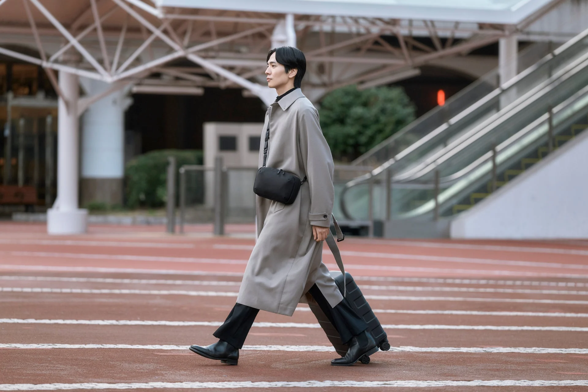 トレンチコートを着た男性がショルダーバッグを肩にかけ、スーツケースを引きながら屋外を歩く都市の風景