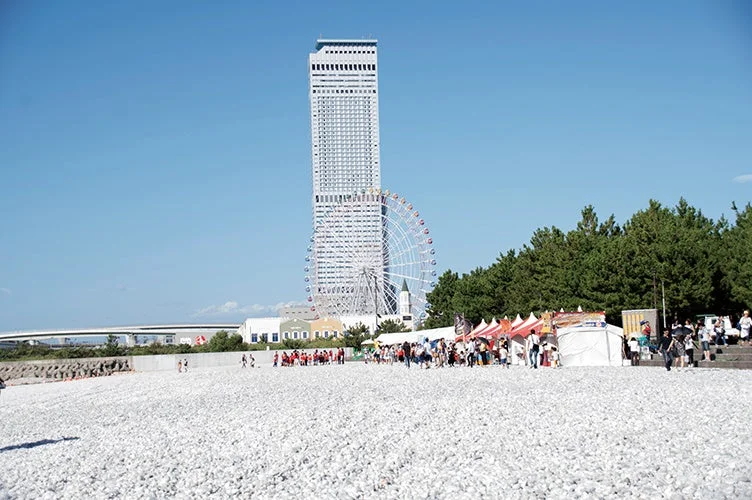 白い小石で覆われた広い屋外スペースでイベントが開催されています。背景には高層ビルと観覧車、そして緑豊かな木々が並び、多くの人々がテントや屋台の周りに集まっています。