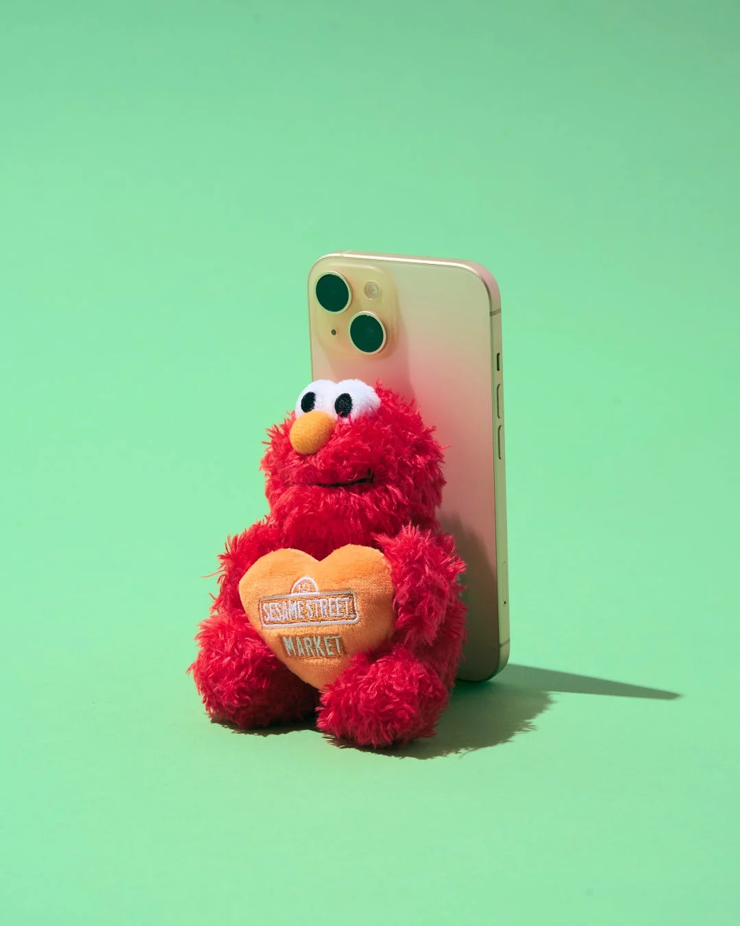 エルモのぬいぐるみがiPhoneにもたれかかり、セサミストリートマーケットと書かれたハートを抱えている様子