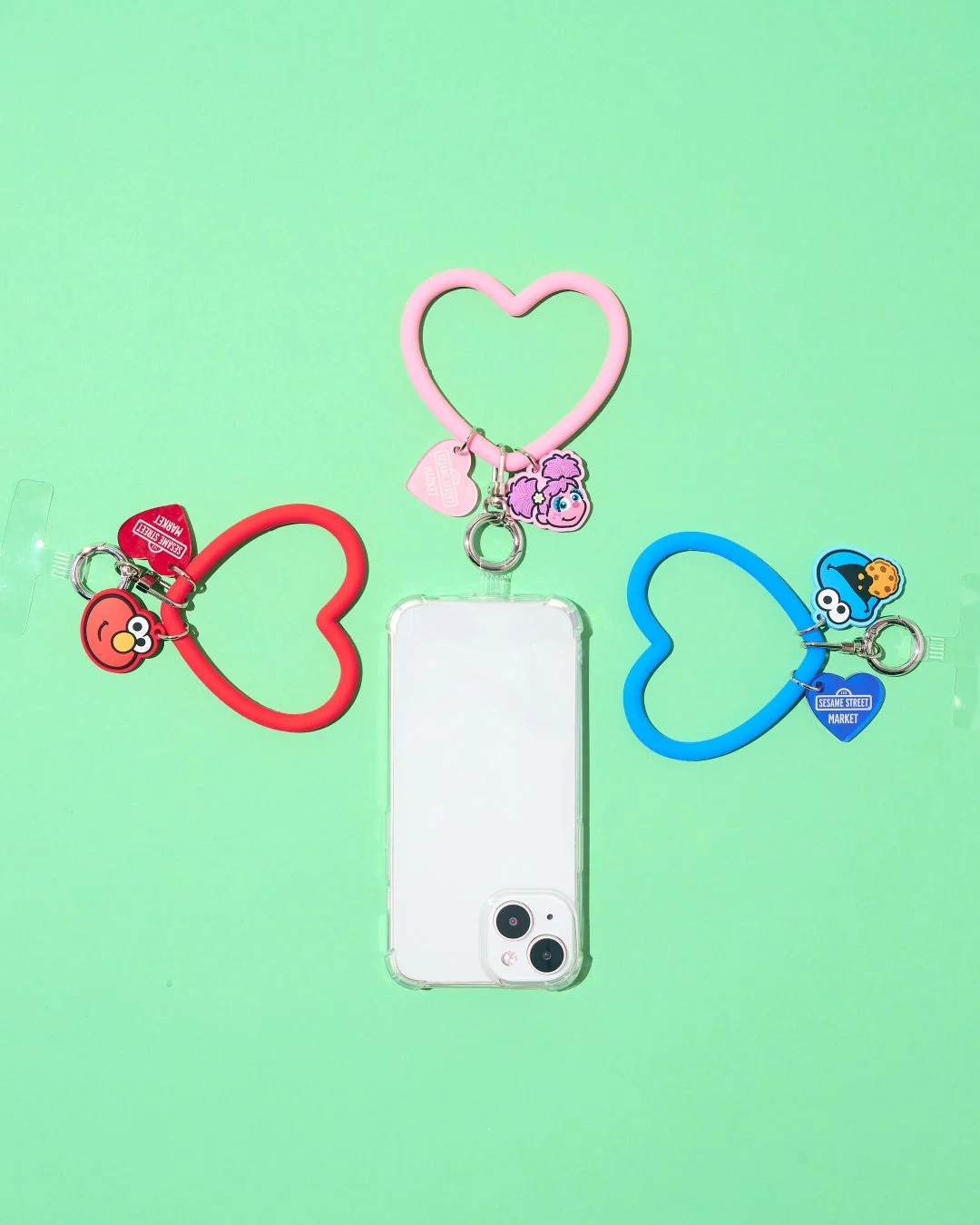 白いスマートフォンとセサミストリートのキャラクター（エルモ、アビー、クッキーモンスター）が付いたハート型スマホストラップ3種