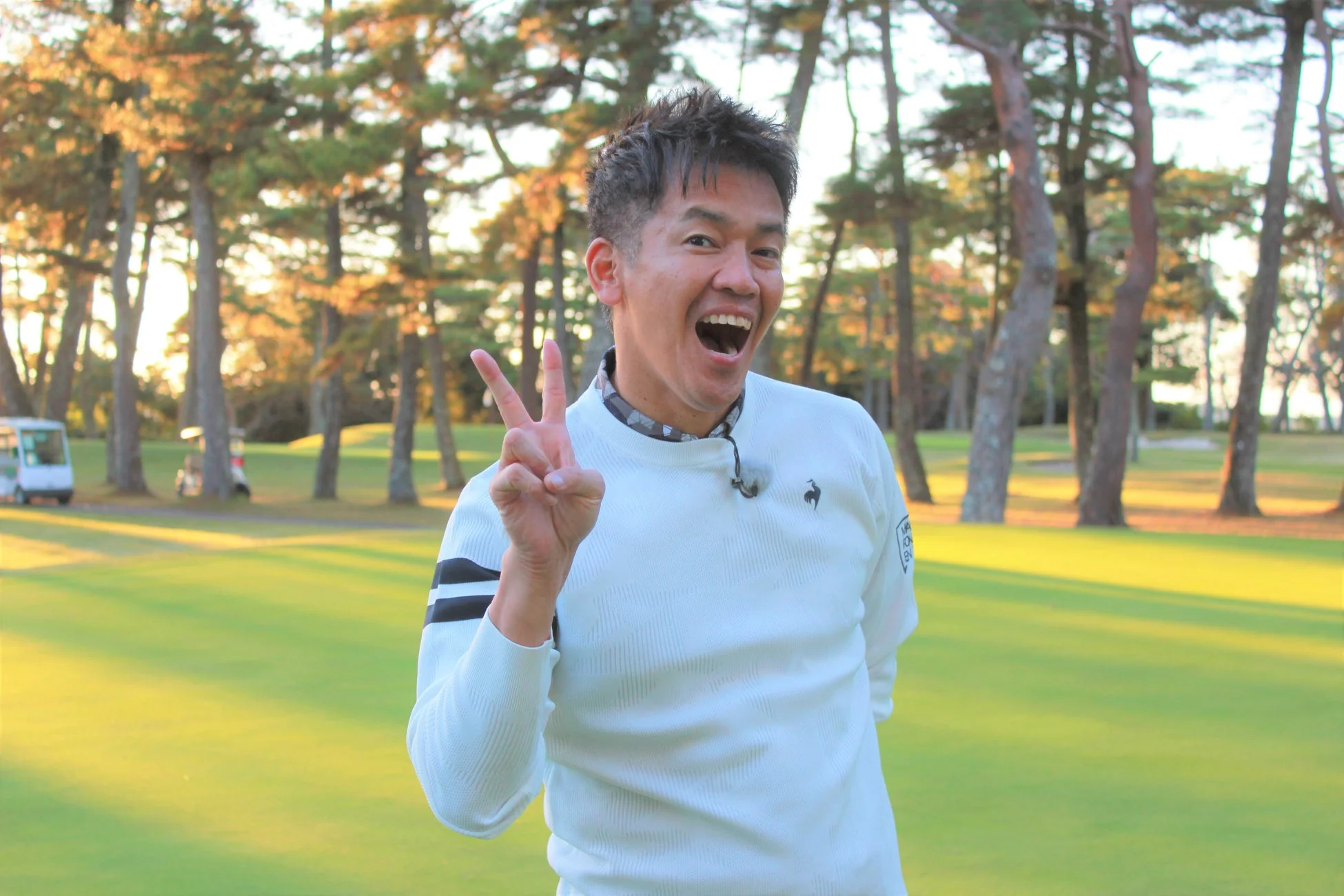 ゴルフ場で白いセーターを着た男性が、満面の笑みでピースサインをしている