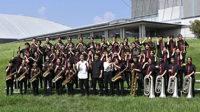 屋外で楽器を持った大勢の学生たちが、黒いシャツと赤いネクタイの制服を着て集合写真を撮っている
