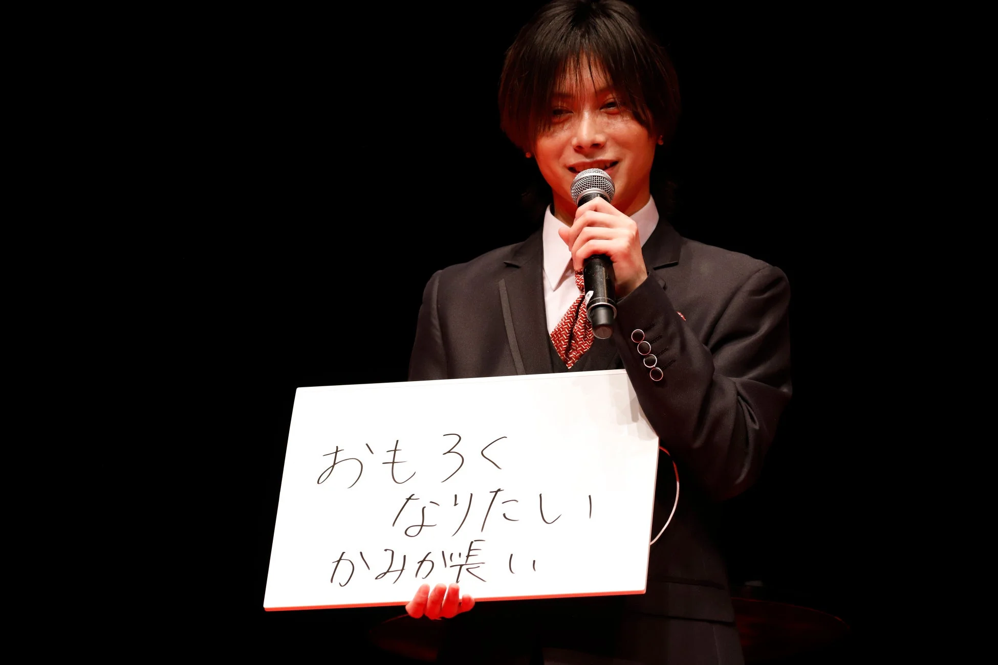 スーツを着た男性がマイクを持ち、ホワイトボードに「おもしろくなりたい かみが長い」というメッセージを掲げ、笑顔で話している様子です。イベントでの発表シーンと見られます。