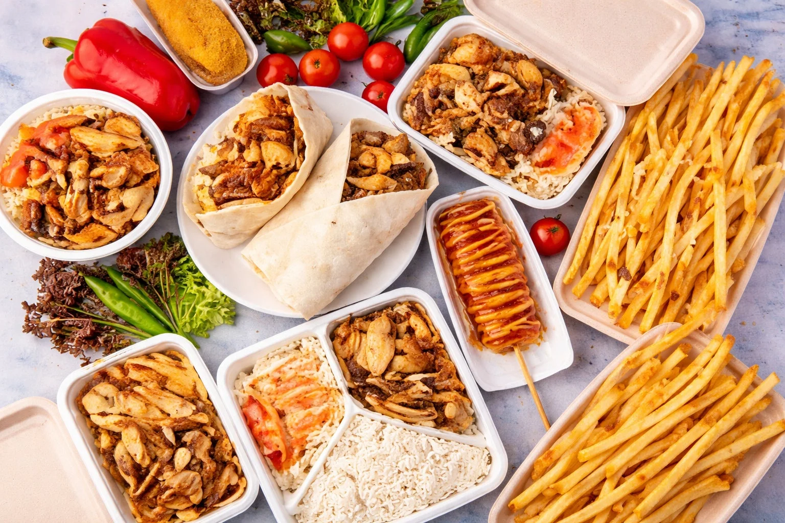 鶏肉とご飯の弁当、ラップサンド、コーン犬、フライドポテトなど、様々なテイクアウト料理がテーブルに並べられた画像