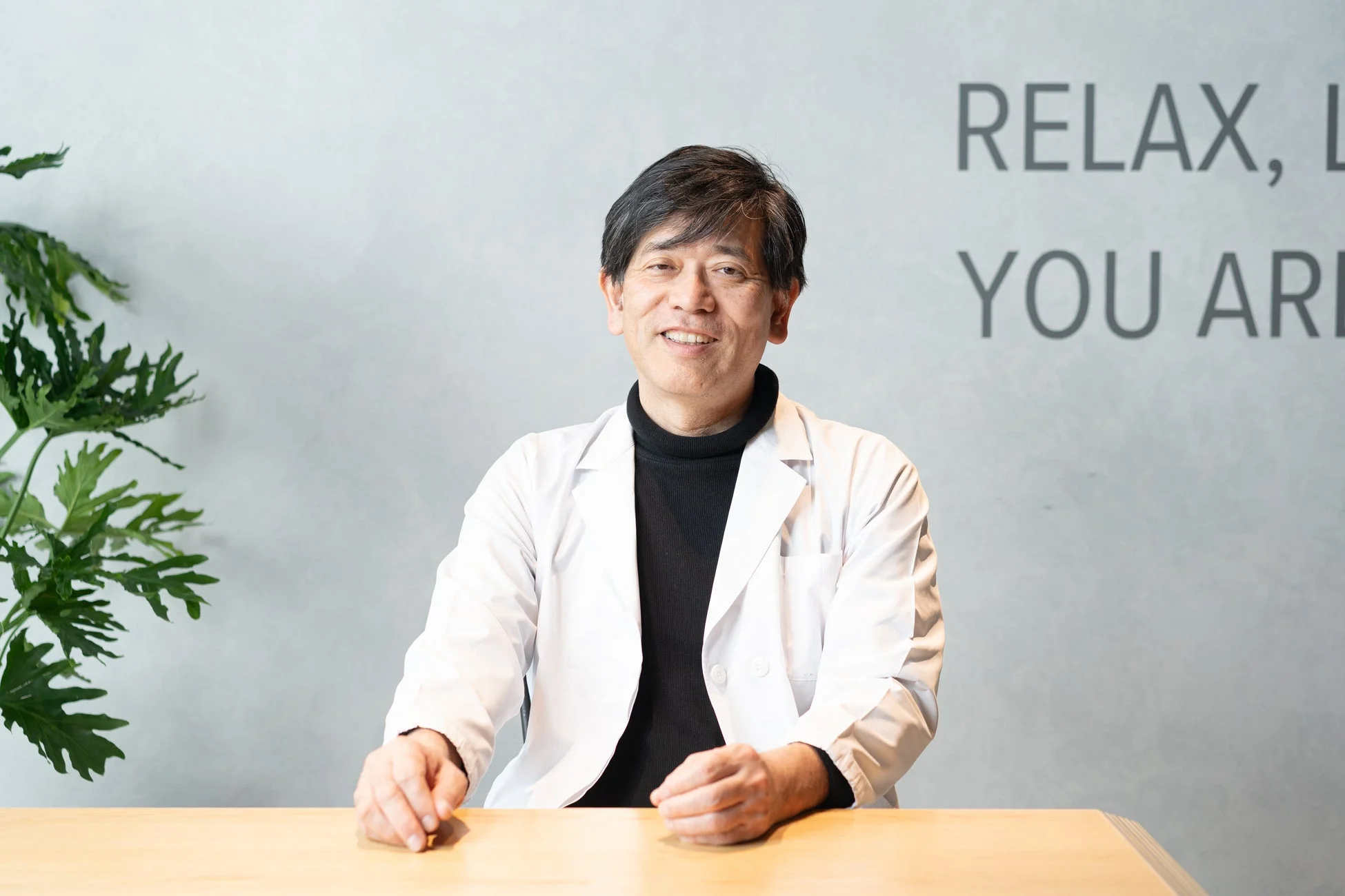 白衣と黒いタートルネックを着用した男性が、笑顔でデスクに座っている写真