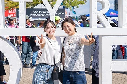 屋外イベントで、白い大きなオブジェの前で笑顔でピースサインをする二人の女性が写っています。背景には多くの人々が見えます。