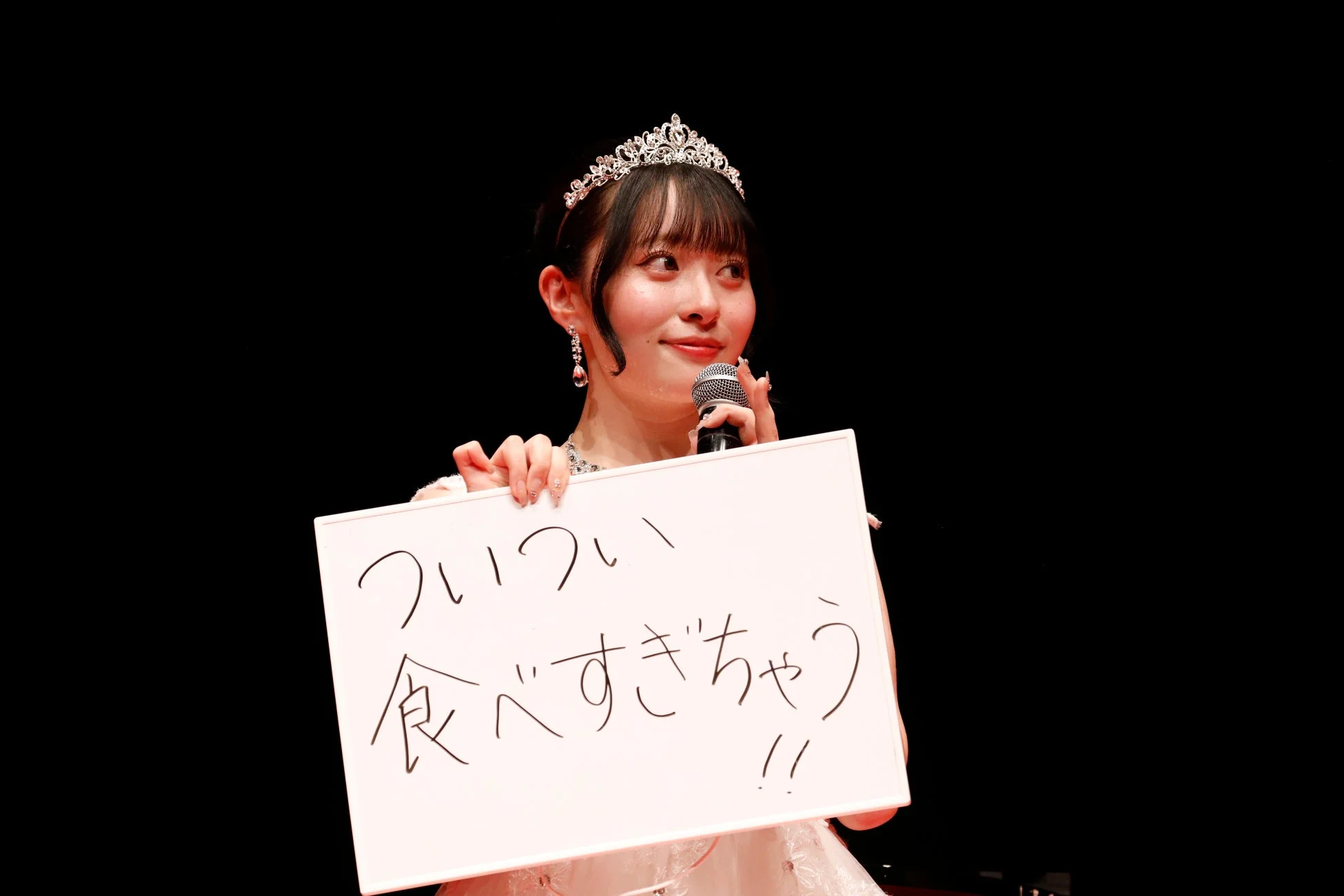 ティアラとドレスを身につけた若い女性がステージ上でマイクを持ち、手書きメッセージ「ついつい食べすぎちゃう!!」と書かれたホワイトボードを掲げています。