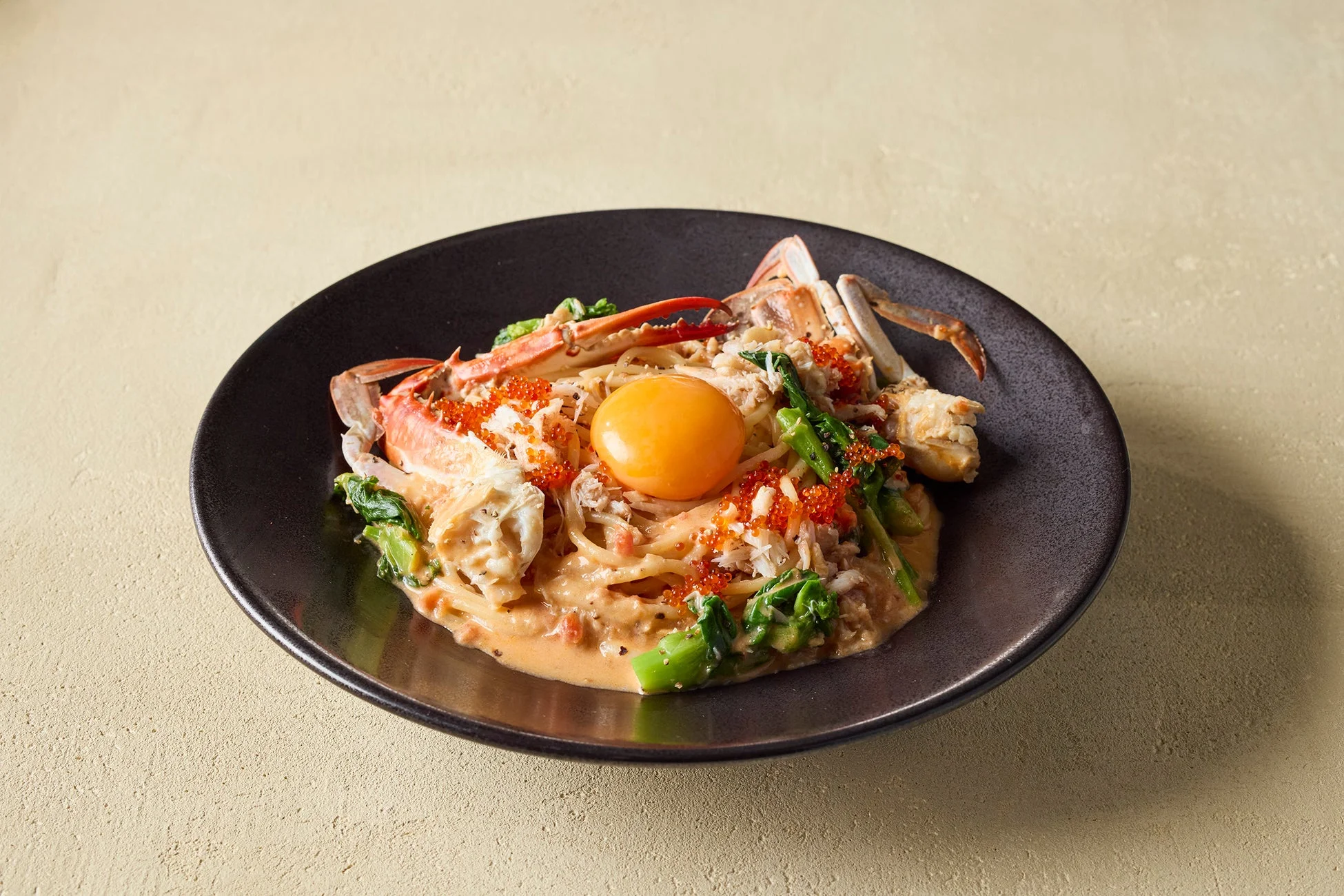 カニ肉とカニの脚、緑の野菜、赤い魚卵、そして鮮やかな生卵の黄身がトッピングされたクリーミーなパスタ料理