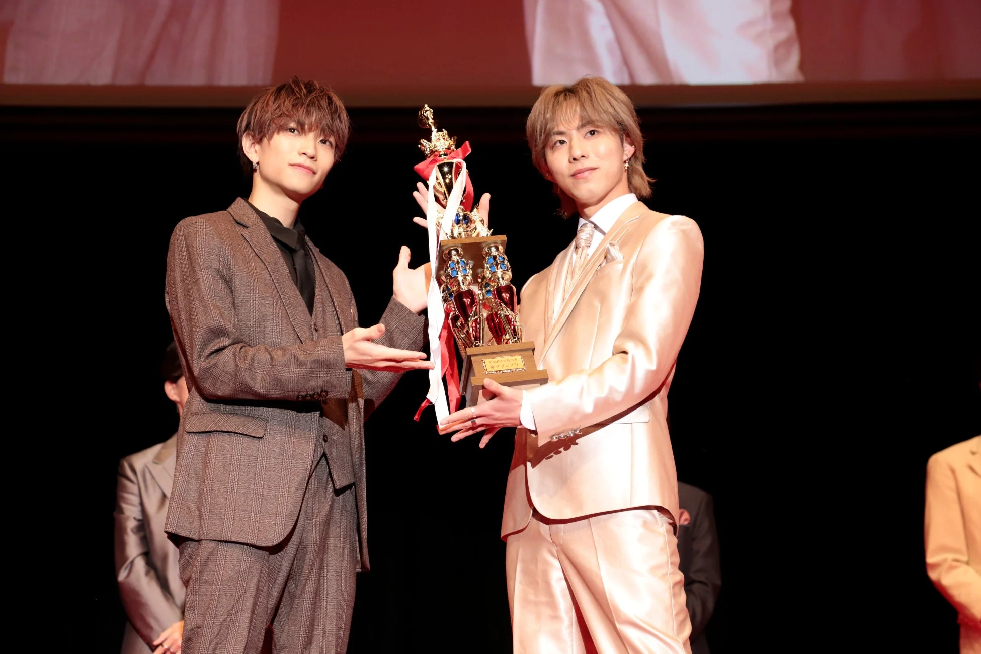 授賞式で光沢のあるスーツを着た2人の若い男性が、誇らしげに「CAMPUS BOYS グランプリ」のトロフィーを掲げている様子。