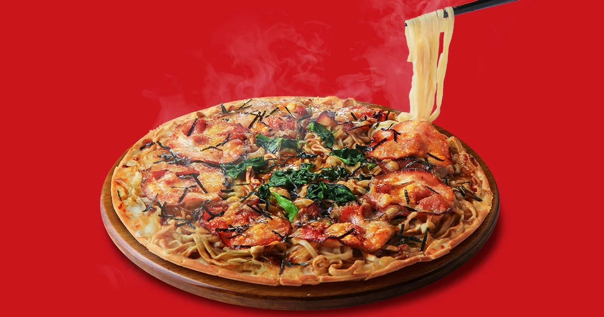 赤い背景に湯気が立つ、麺をベースにしたユニークな和風ピザが木製の台に乗っています。箸で麺が持ち上げられており、豚肉やほうれん草がトッピングされた、食欲をそそる一品です。