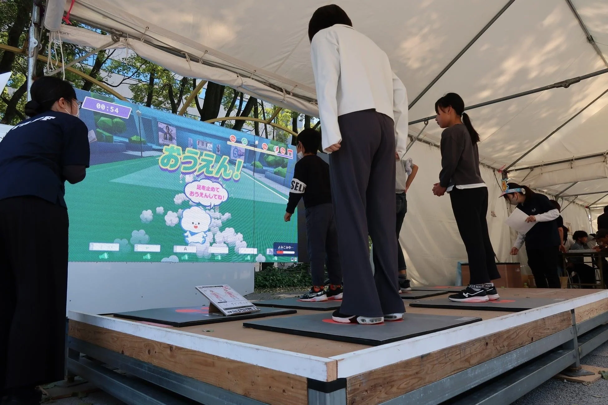 屋外イベントの白いテント内で、人々が大型スクリーンに映るインタラクティブゲームに参加しています