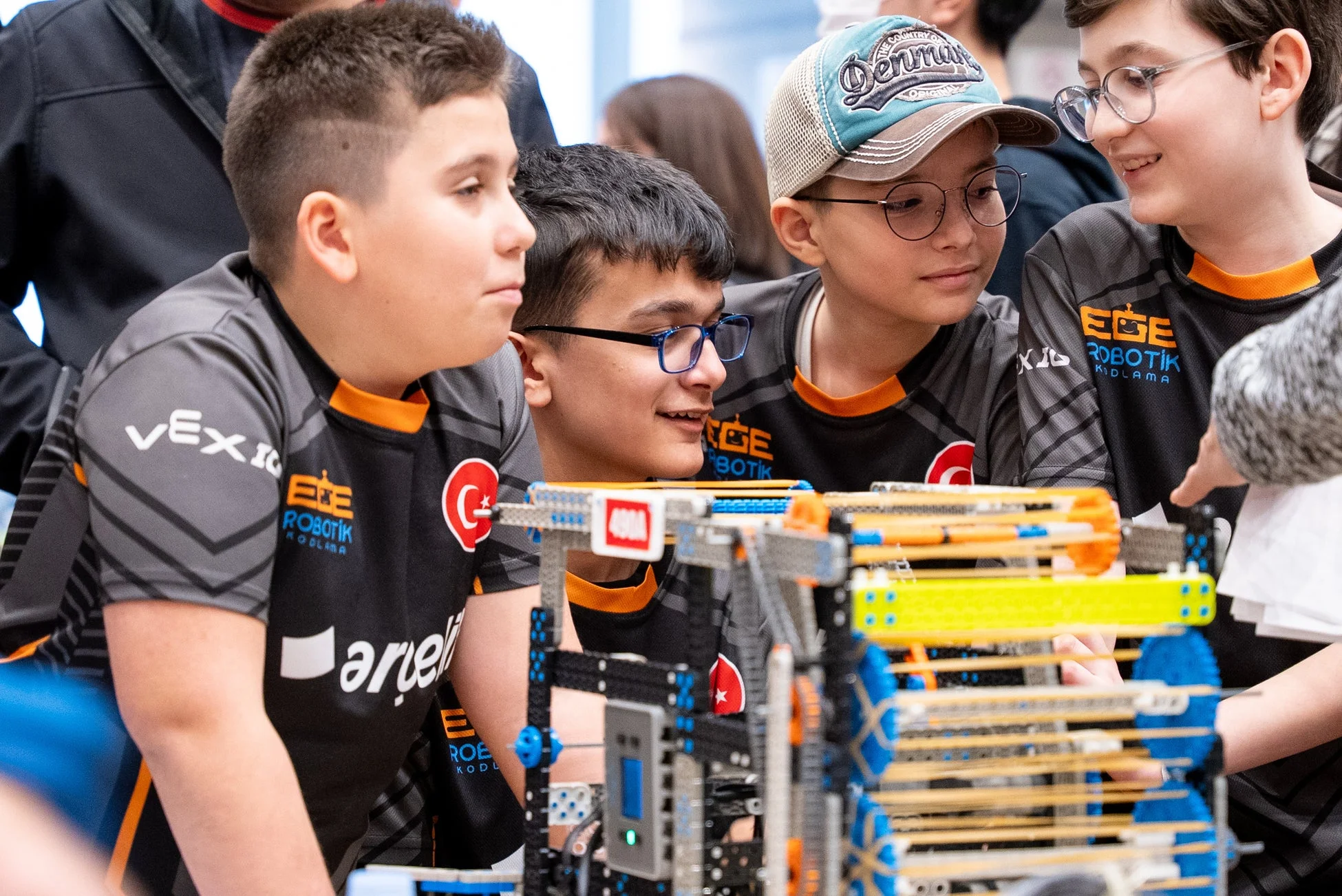 VEX IQとEGE ROBOTIKのロゴ入りシャツを着た少年たちが、ロボット競技会で複雑なロボットに集中し、協力し合っている様子。