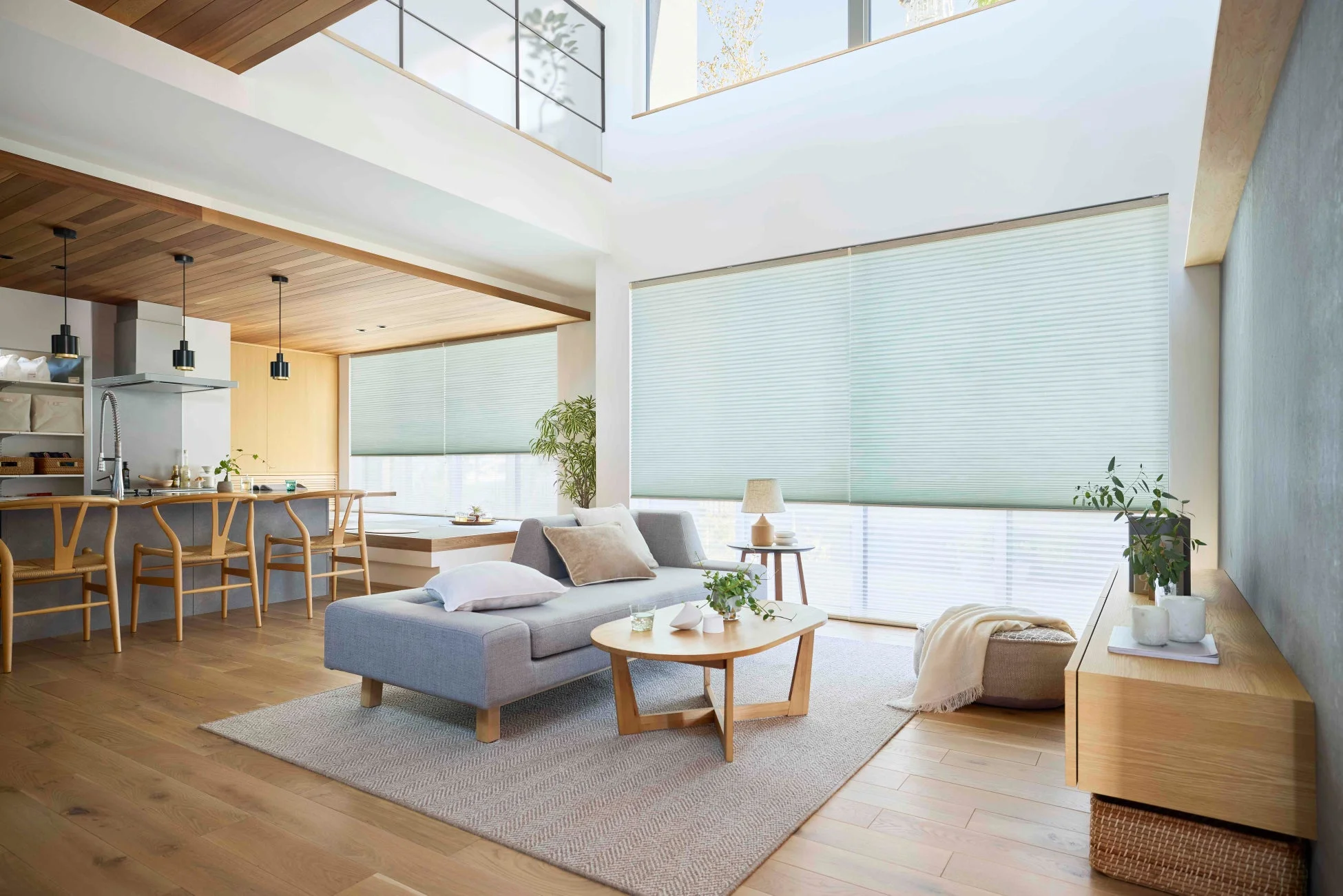 広々としたリビングダイニングキッチンが一体となった、明るく開放的なモダンな住宅空間です。木目調の床と家具、白い壁が基調で、大きな窓にはプリーツスクリーンが設置されています。シンプルなグレーのソファやおしゃれな照明が配置され、ナチュラルで洗練された印象を与えます。
