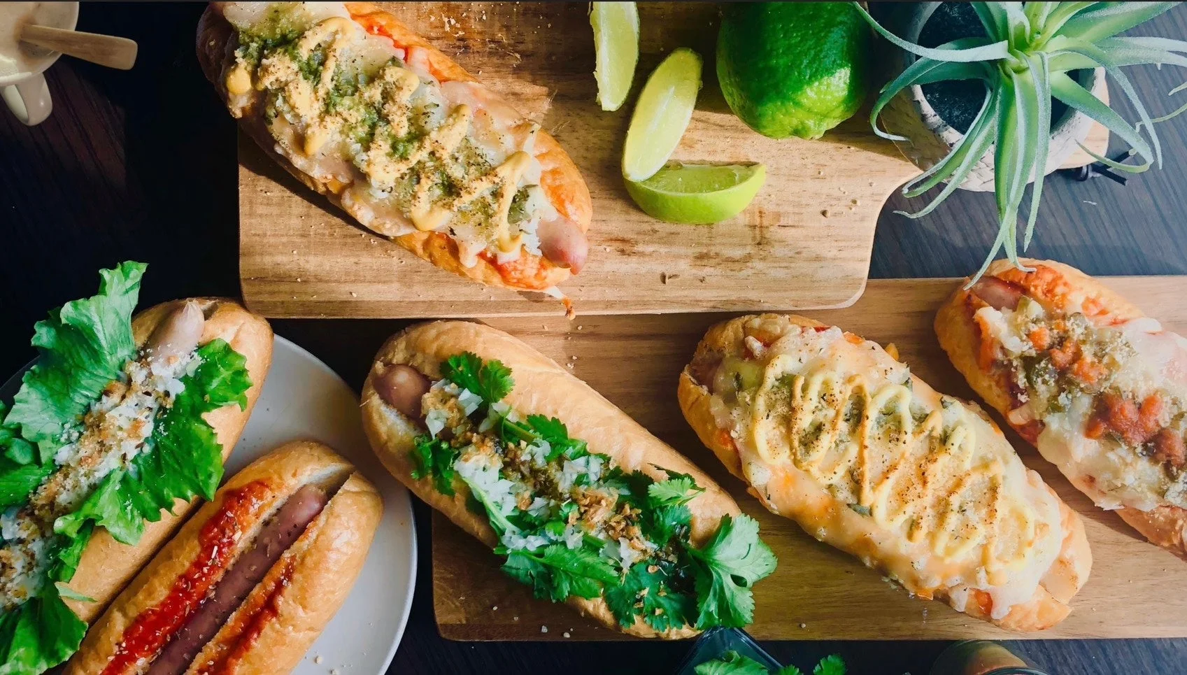 様々なトッピングが施されたホットドッグが複数、木製のボードと白い皿に盛り付けられた画像