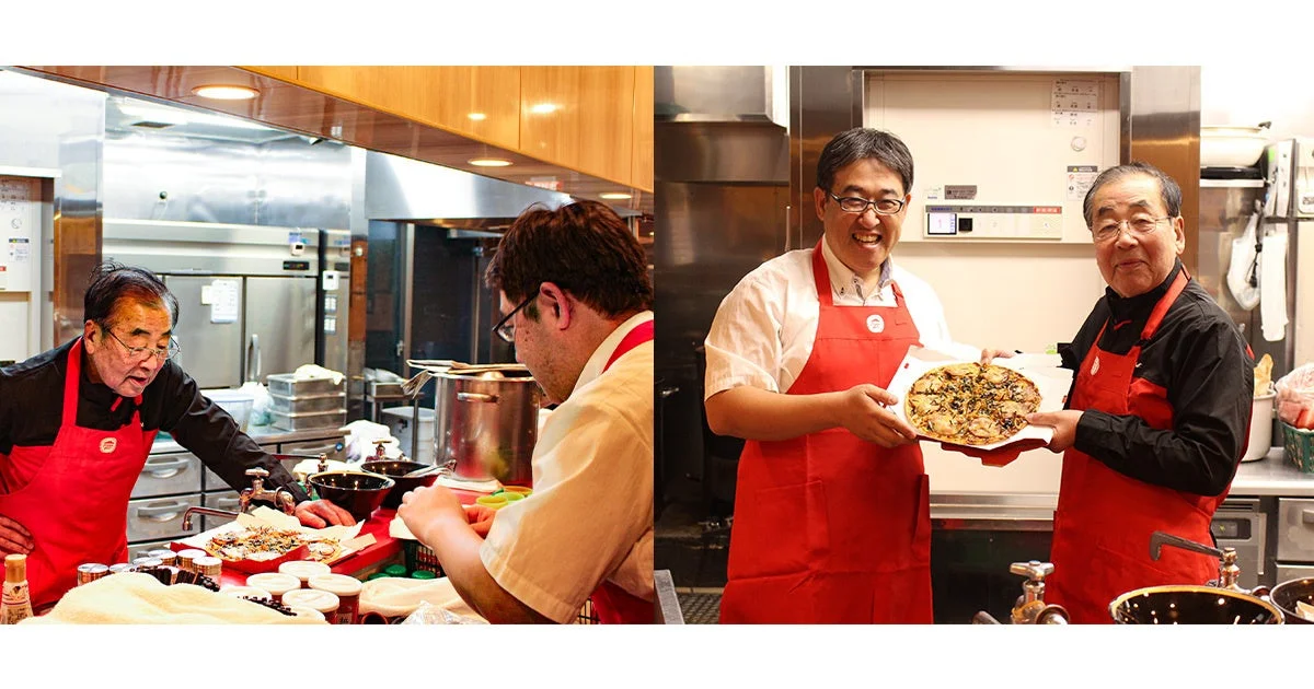 赤いエプロンを着た2人の男性が厨房で料理を準備し、完成したピザのような料理を笑顔で披露している様子が捉えられています。