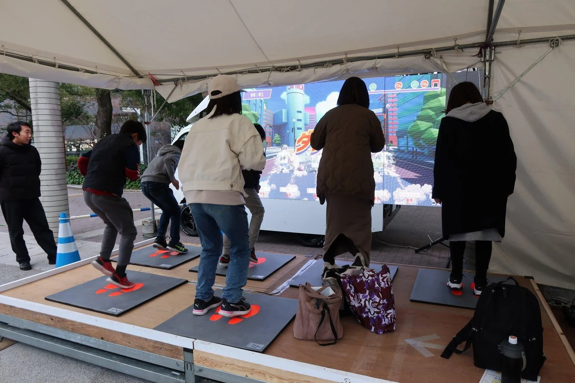 屋外のテントの下で、数人が大型デジタルスクリーンに映し出されたインタラクティブゲームを体験しています