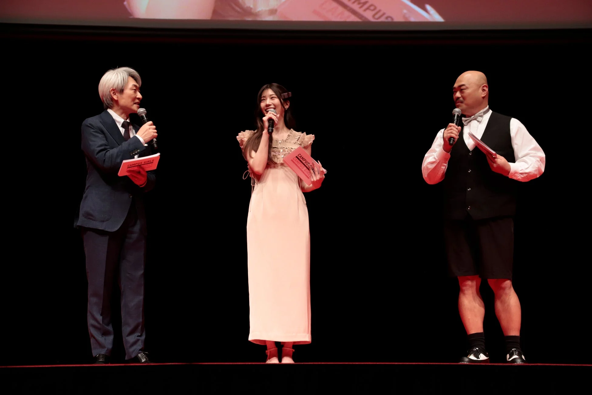 暗いステージ上で、中央に笑顔の女性、左右に男性がそれぞれマイクを持って立っています。3人は何かを話している様子で、手には「CAMPUS GIRLS」と書かれた資料を携えています。