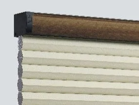 画像は、ブラウンのヘッドレールが付いたクリーム色のプリーツブラインドのクローズアップです。ブラインドはアコーディオン状に折りたたまれており、生地の質感が見て取れます。窓辺のインテリアを想起させます。