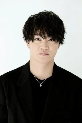 白い背景をバックに、黒いジャケットとTシャツを着用し、銀色のネックレスをつけた黒髪の男性がカメラをまっすぐ見つめているポートレートです