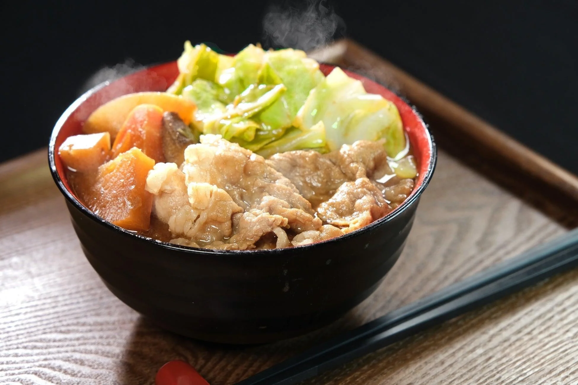 湯気を立てる温かい肉と野菜の煮込み料理が黒い器に盛られています