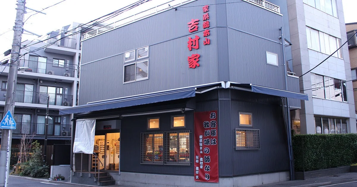 家系総本山として知られる「吉村家」の店舗外観を捉えた一枚。グレーの建物に赤い文字の店名と、「お客様は我が味の師なり」と書かれた赤い垂れ幕が目を引きます。