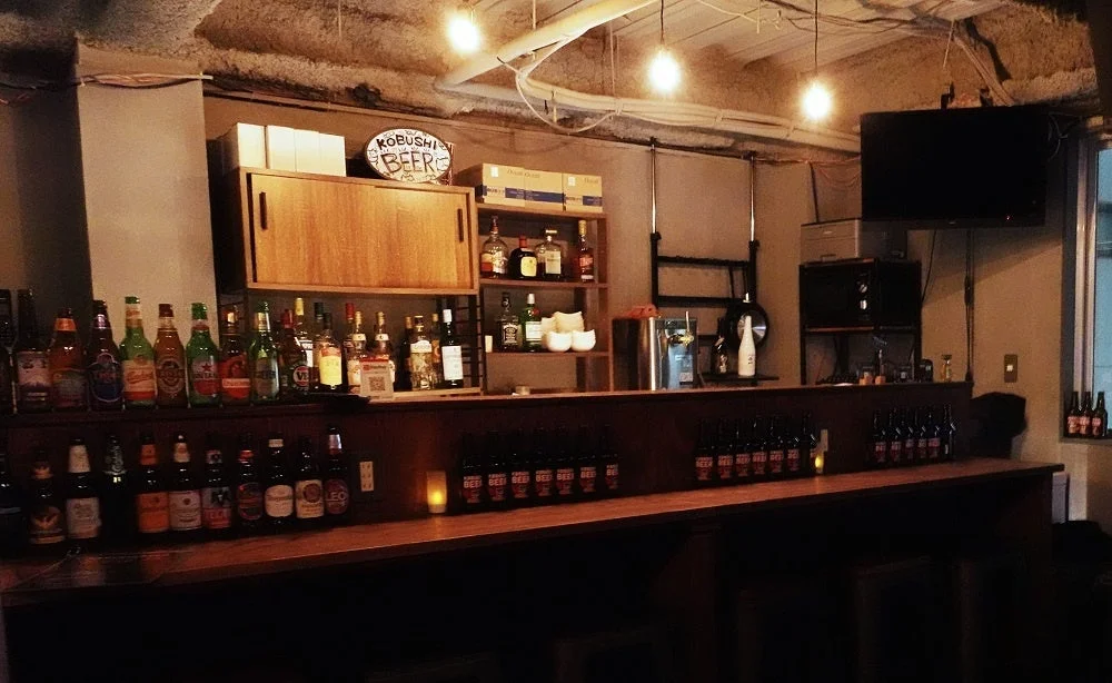 薄暗い照明のバーの店内風景。木製のカウンターには様々な種類のビールや洋酒のボトルが並べられ、上部にはKOBUSHI BEERの看板が見える。