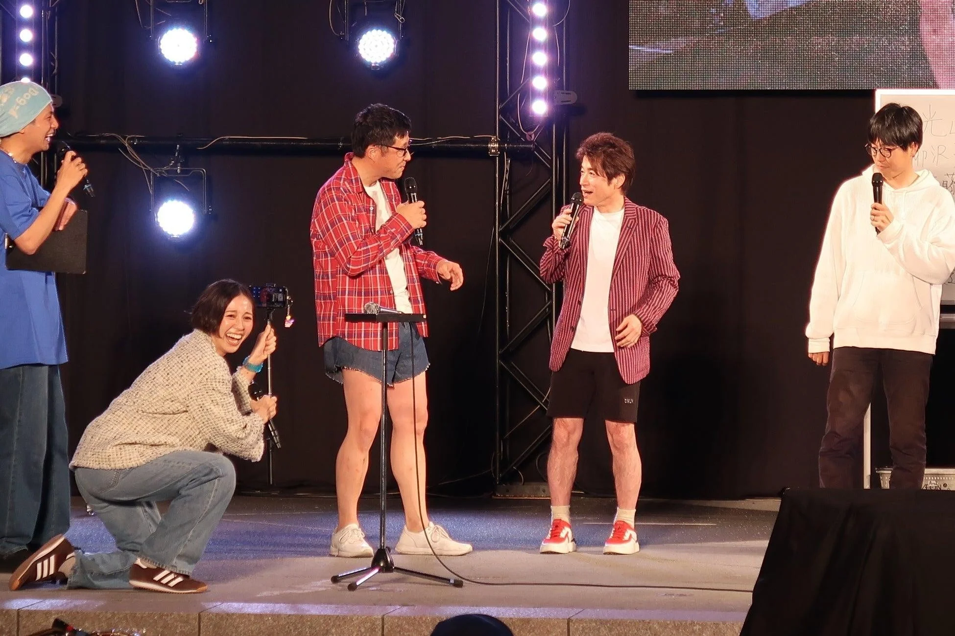 ステージ上で数人の男性がマイクを手にトークを繰り広げ、一人の女性がカメラでその様子を撮影しながら楽しそうに笑っている