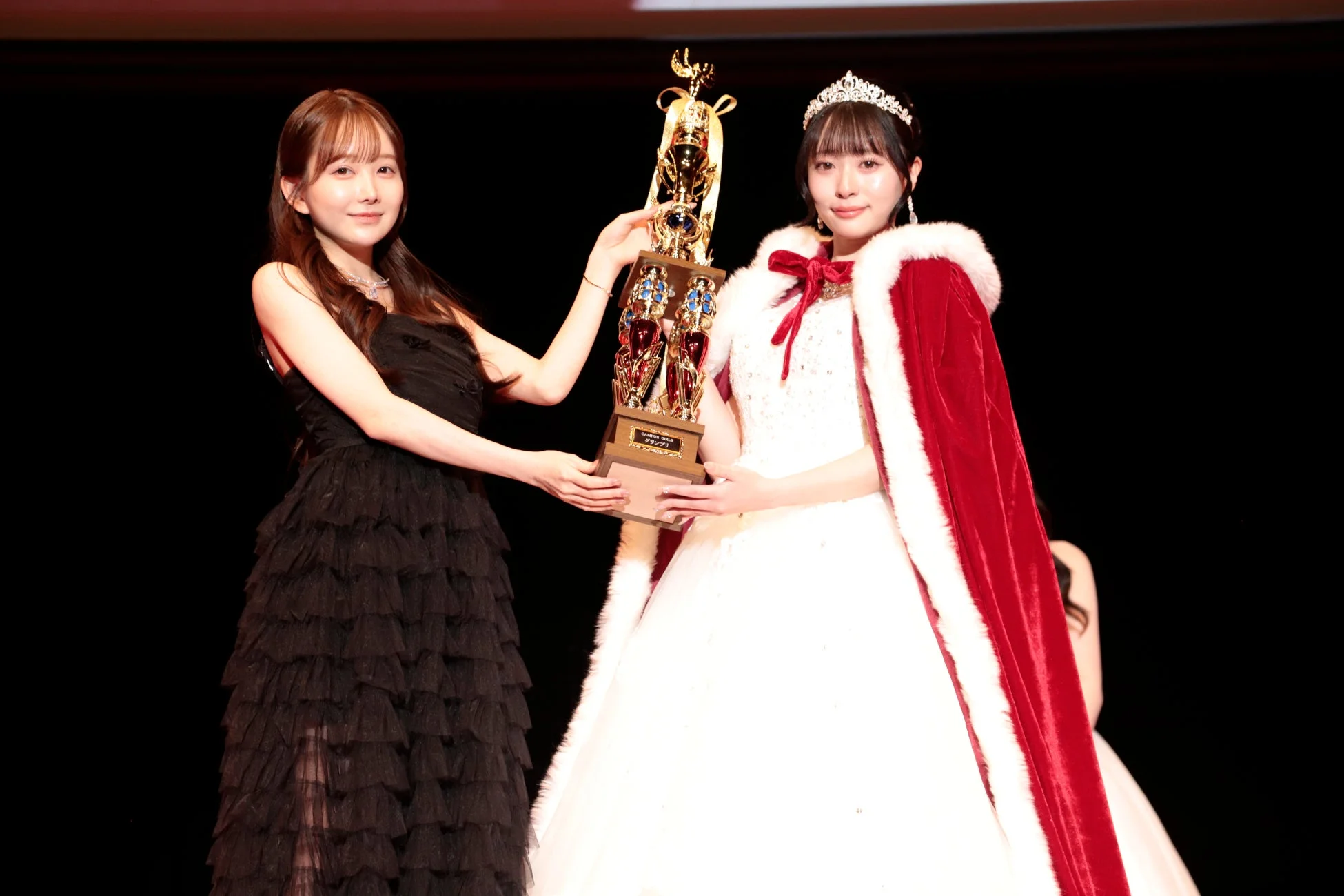 ステージ上で、黒いドレスの女性が白いドレスと赤いケープをまとった女性に大きなトロフィーを授与しています。ミスコンテストのような華やかな表彰式の様子です。