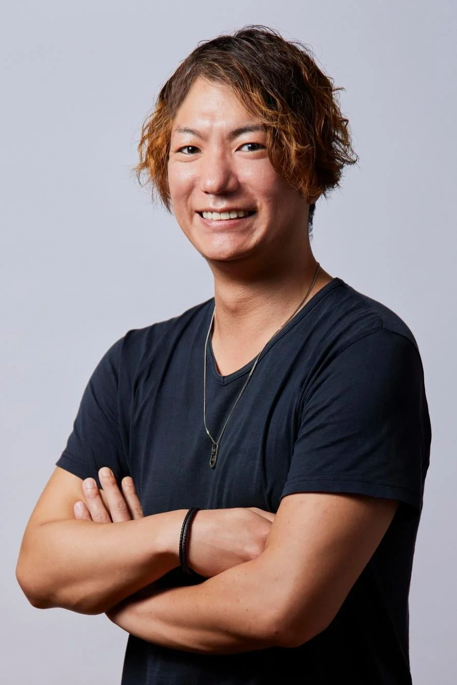 明るい茶色の髪をした若い男性が、黒いTシャツを着て腕を組み、カメラに向かって笑顔を見せているポートレート