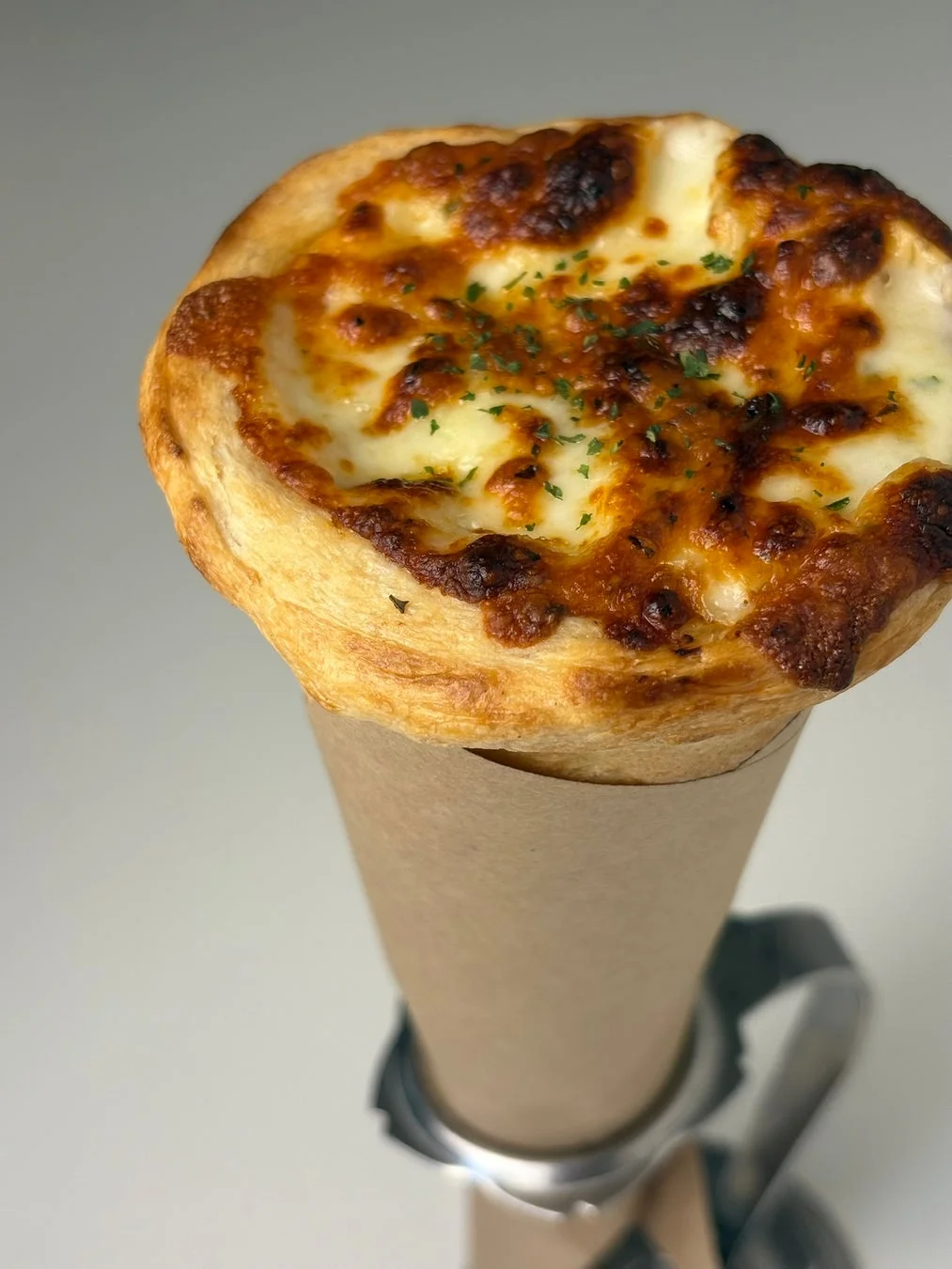 こんがりと焼けたチーズとパセリが乗ったパイが、茶色の紙コーンに入った画像