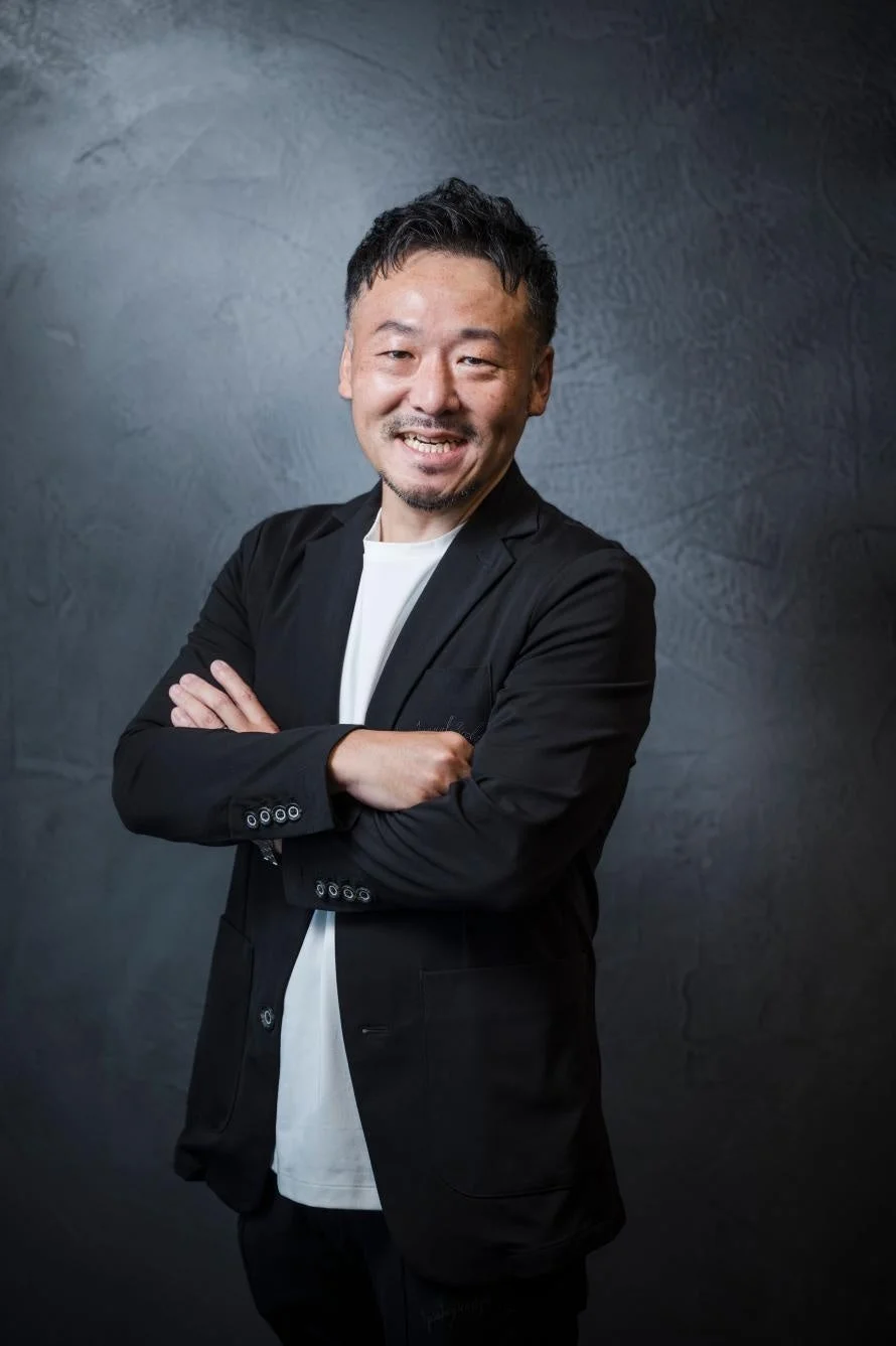 黒いジャケットと白いTシャツを着用し、腕を組んで笑顔を見せる中高年の日本人男性