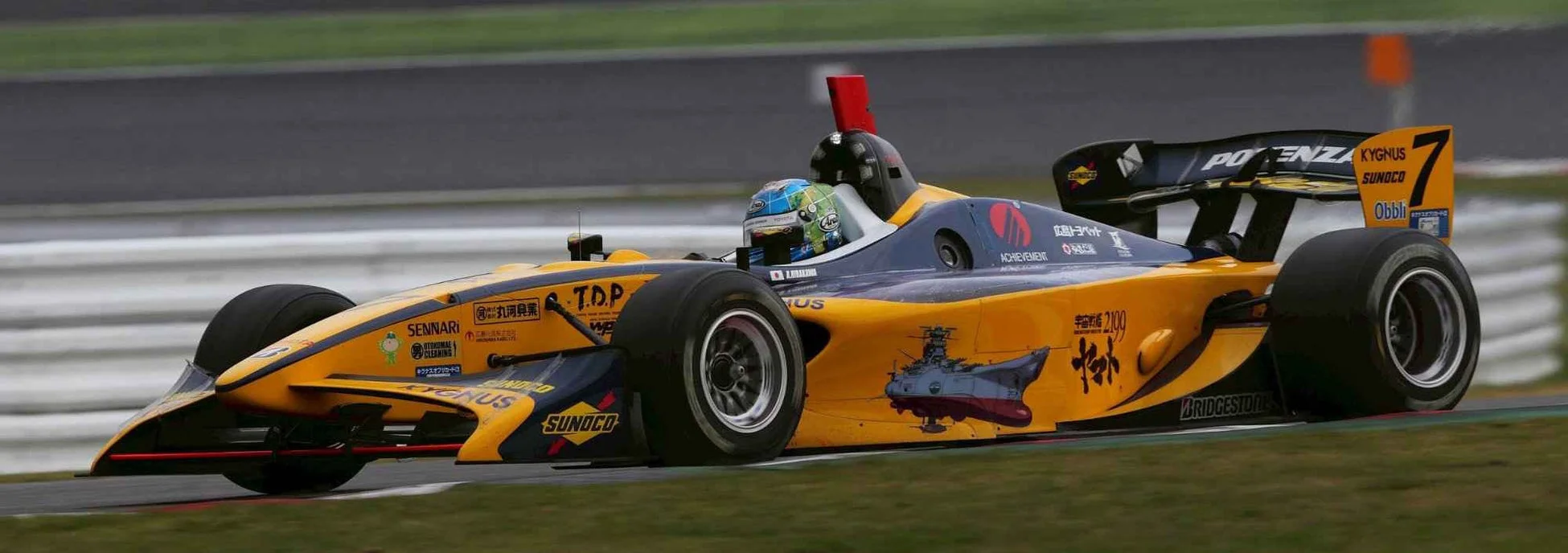 アニメ「宇宙戦艦ヤマト2199」のラッピングが施されたフォーミュラカーが、サーキットを走行している様子を捉えた一枚。ドライバーも搭乗しており、多くのスポンサーロゴが見える。