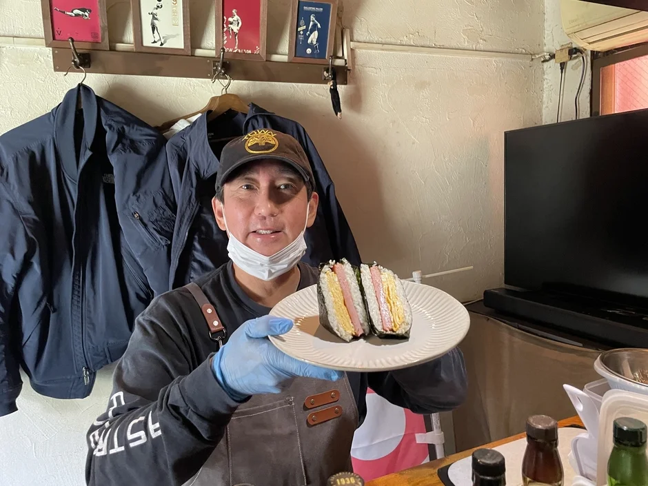 マスクとエプロンを着用した男性が、スパムと卵焼きを挟んだおにぎりを2つ載せた白い皿を差し出しています