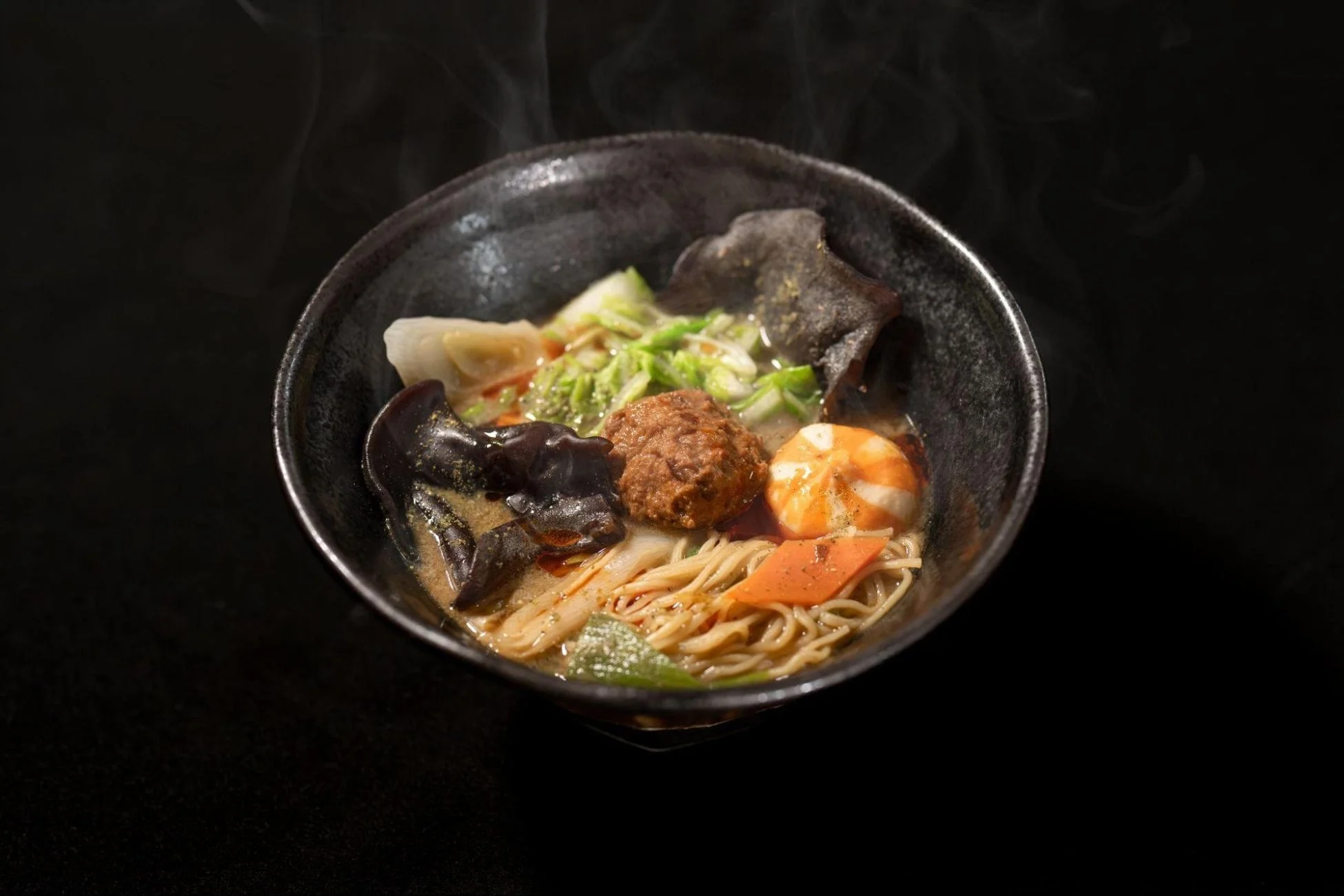 黒い器に盛られた温かい麺料理で、肉団子、エビ、キクラゲ、白菜、人参などの具材が豊富に入っており、湯気が立ち上る様子が食欲をそそります