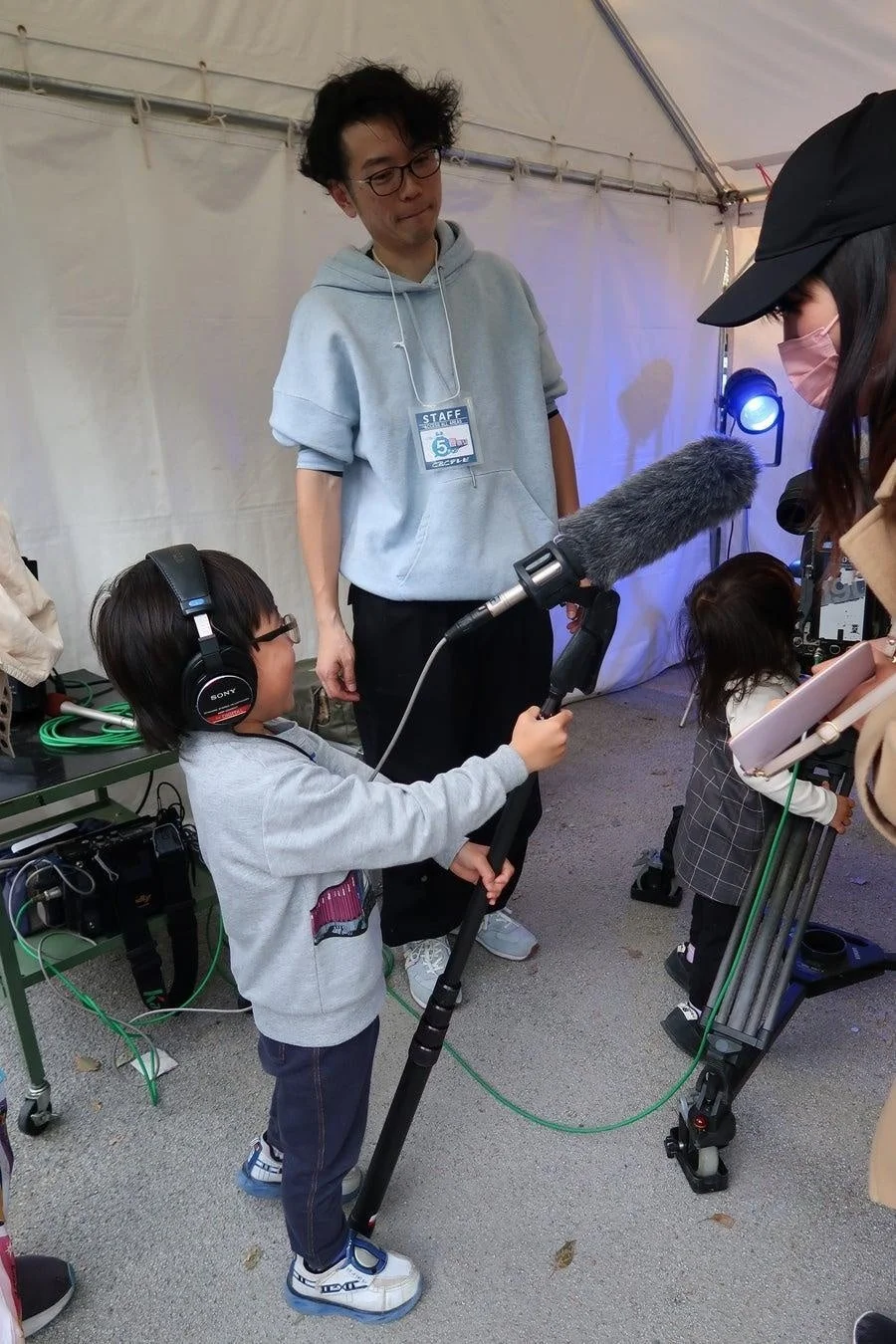 子供がブームマイクとヘッドホンを装着し、大人のスタッフに囲まれながら撮影体験をしている様子