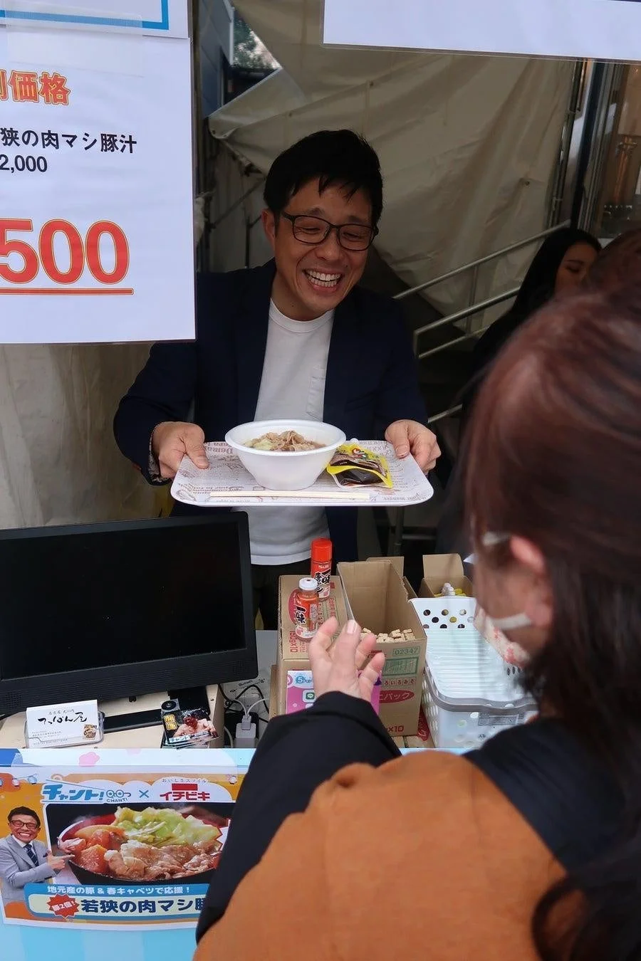 笑顔の男性が、屋外の屋台で客に「若狭の肉マシ豚汁」を手渡しています